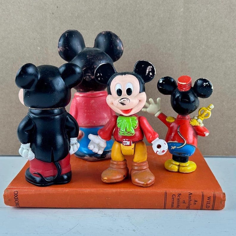 May include: Four vintage Mickey Mouse figurines in various poses, standing on a vintage book. The figures are in red, black, yellow, and white. One figure is waving, and another is dressed as a band leader.