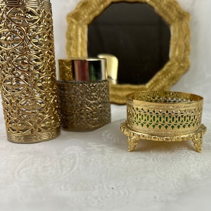 May include: A collection of vintage gold-toned vanity items. Includes a perfume bottle with an openwork design, a small jar, and a decorative tray with a green interior. A gold-framed mirror is in the background.