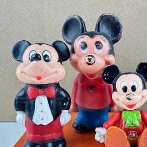 May include: Three vintage Mickey Mouse figurines. The figurines are in various poses and outfits, including a tuxedo, a red shirt, and blue shorts. The figures have black ears, red, white, and black clothing, and are made of a hard plastic material.