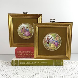 May include: Two framed vintage porcelain plaques with gold frames. Each plaque features a circular porcelain scene of people in period clothing. The frames are square with a dark green background. The plaques are displayed on top of books.