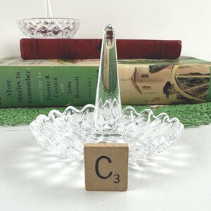K&ouml;nnte beinhalten: Ein Ringhalter aus klarem Glas mit einem zentralen Kegel f&uuml;r Ringe und einem gewellten Sockel mit Rautenmuster. Ein kleines quadratisches Holzpl&auml;ttchen mit dem Buchstaben C und der Zahl 3 befindet sich vor dem Sockel des Glasringhalters.