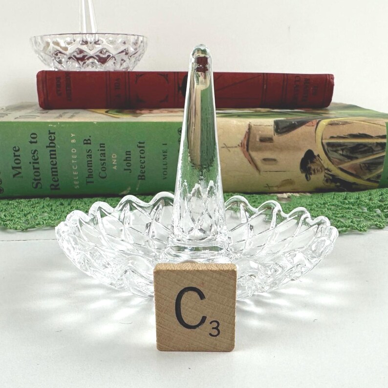K&ouml;nnte beinhalten: Ein Ringhalter aus klarem Glas mit einem zentralen Kegel f&uuml;r Ringe und einem gewellten Sockel mit Rautenmuster. Ein kleines quadratisches Holzpl&auml;ttchen mit dem Buchstaben C und der Zahl 3 befindet sich vor dem Sockel des Glasringhalters.