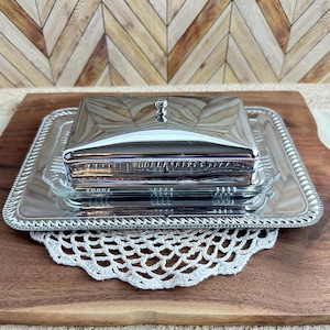 May include: A rectangular silver-toned metal butter dish with a matching lid featuring a small knob handle. The dish sits on a rectangular silver-toned metal tray with a decorative rope-style border edge and a clear glass insert.