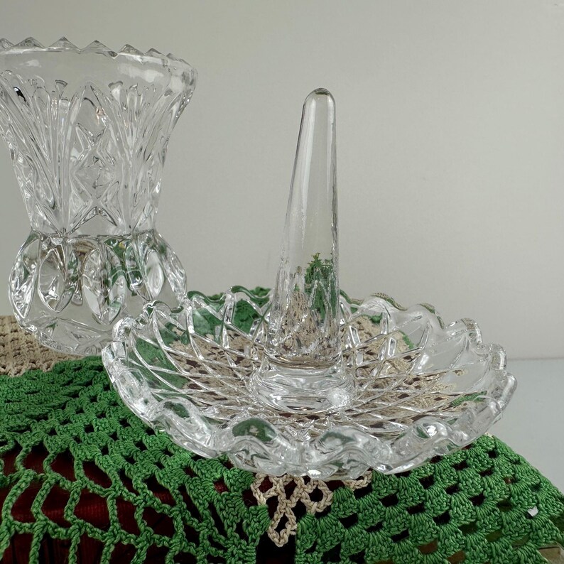K&ouml;nnte beinhalten: Ein Ringhalter aus klarem Glas mit einem zentralen konischen Stift auf einem gewellten Sockel mit Rautenmuster, begleitet von einer passenden Vase aus klarem Glas mit ausgestelltem Rand und vertikalem Schliffdekor.