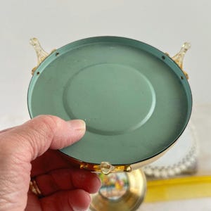 May include: A vintage trinket box with a light green interior and a gold-colored exterior. The box has a round shape with decorative clear plastic accents and a small, colorful image on the bottom.