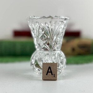 K&ouml;nnte beinhalten: Eine Vase aus klarem Glas mit einem facettierten Pressmuster, scharfen geometrischen Schliffen und einem ausgestellten Rand. Ein kleines, quadratisches Holzpl&auml;ttchen mit dem aufgedruckten Buchstaben A und der Zahl 1 steht vor der Basis.