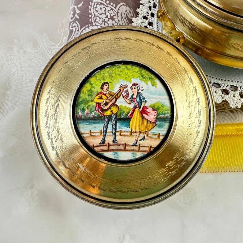 May include: A round, gold-toned trinket box with a painted scene of two figures on a bridge, one playing a guitar. The figures are dressed in colorful period clothing. The box has a decorative, textured edge.