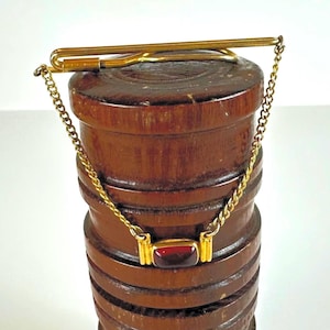 May include: A gold-toned tie clip with a chain and a rectangular, red gemstone. The clip is attached to a wooden, cylindrical object with a series of horizontal grooves. The chain hangs down from the clip, connecting to the gemstone.