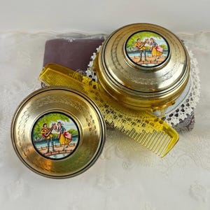 May include: Two vintage gold-toned compacts and a yellow comb. Each compact features a colorful painted scene of a couple with a guitar on a bridge. The comb is a translucent yellow color.