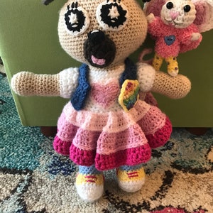May include: Crocheted pug doll with a pink and white dress, blue vest, and rainbow patch. The pug has large black and white eyes, a black nose, and small ears. A crocheted mouse doll with pink ears and a blue heart is next to the pug.