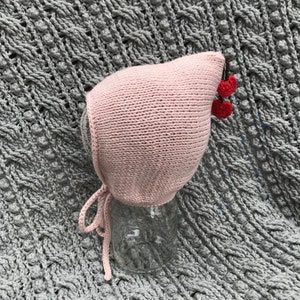 May include: A light pink knitted baby bonnet with a cherry accent. The bonnet has a rounded shape with ties and is decorated with two red cherries. The bonnet is displayed on a glass jar, set against a textured gray background.