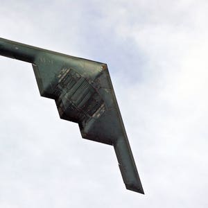 May include: A dark gray stealth bomber aircraft flying in a clear blue sky with white clouds.