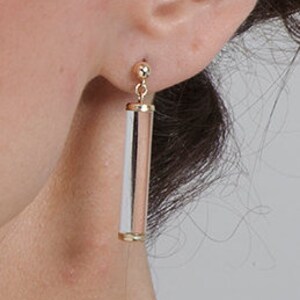 May include: A pair of gold and white dangle earrings with a cylindrical design. The earrings are made of metal and have a polished finish.