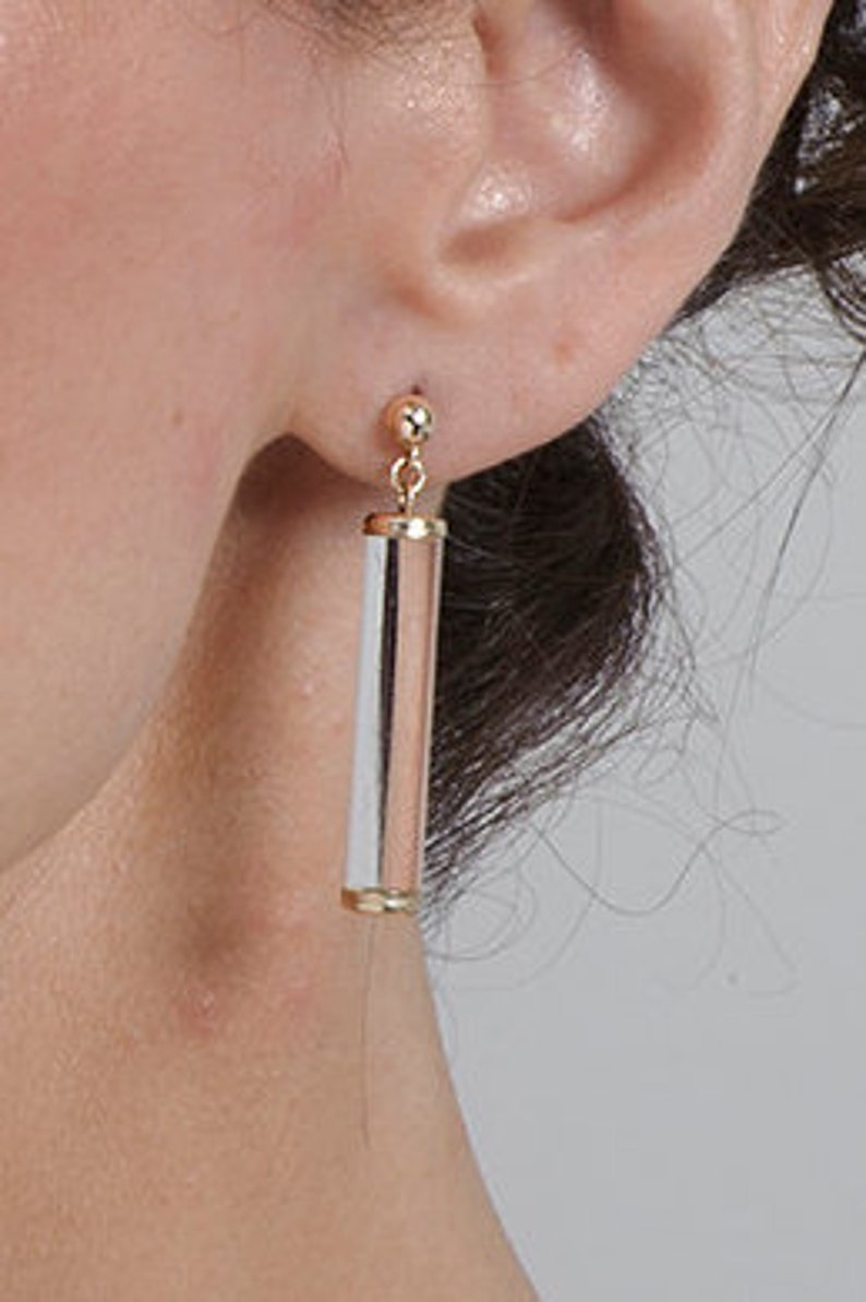 May include: A pair of gold and white dangle earrings with a cylindrical design. The earrings are made of metal and have a polished finish.