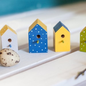 Può includere: Quattro casette per uccelli in legno in miniatura dipinte in diversi colori e motivi. Le casette per uccelli sono disposte in fila su una superficie bianca. Le casette per uccelli sono dipinte di blu con pois bianchi, giallo con strisce nere, bianco con accenti gialli e verde con pois bianchi.