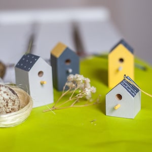 May include: Four miniature wooden birdhouses painted in shades of gray, yellow, and white. The birdhouses have small, round openings for birds to enter. The birdhouses are arranged on a green surface with a small nest and a sprig of dried flowers.