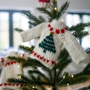 May include: A white knitted Christmas sweater ornament with a green embroidered Christmas tree and red hearts. The ornament is hanging on a green Christmas tree.