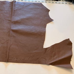May include: A piece of brown leather with a textured surface. A measuring tape is placed along the top edge, showing inch measurements. The leather is cut in an irregular shape, with a white surface in the background.
