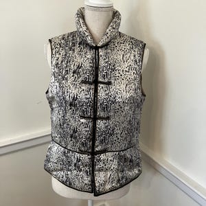 May include: A sleeveless waistcoat featuring a high collar and a black and white speckled animal print pattern. The garment includes a front closure with three horizontal black toggle fasteners and black piping trim along the edges and seams.