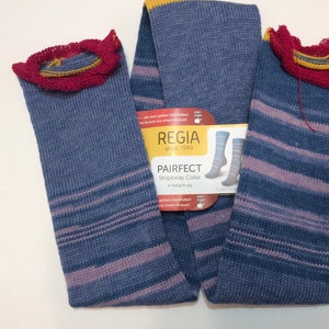 May include: Two pairs of blue and purple striped socks with a yellow and red crocheted trim. The socks are laid out flat on a white surface. A label for Regia Pairfect Stripeway Color yarn is visible in the center of the image.