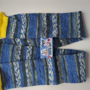 May include: A knitted scarf with blue, gray, and white stripes and yellow ends. The scarf has a tag that reads "Best of Opal". The scarf is made of soft yarn and is likely handmade.