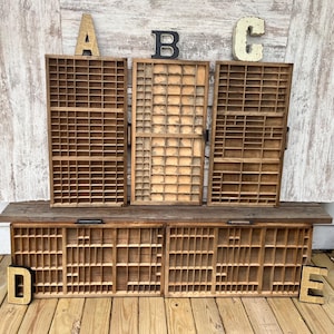 May include: Five rectangular wooden printer trays with numerous small compartments arranged in a grid pattern. The trays are displayed with large, decorative letters A, B, C, D, and E placed on or near them, featuring a natural wood finish.