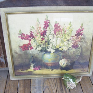 May include: A framed vintage print of a still life featuring a bouquet of pink, white, and red snapdragon flowers in a blue and gold vase. The vase is sitting on a table with a decorative plate and a small teapot.