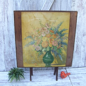 May include: A framed floral still life painting featuring a green vase filled with colorful flowers. The painting has a yellow background and is displayed on a small wooden easel. The frame is a dark brown color.