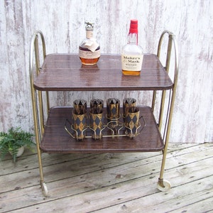 May include: A vintage gold metal bar cart with two brown wooden shelves. The cart has four wheels and is holding a bottle of Maker's Mark whiskey, a bottle of liquor, and six glasses.