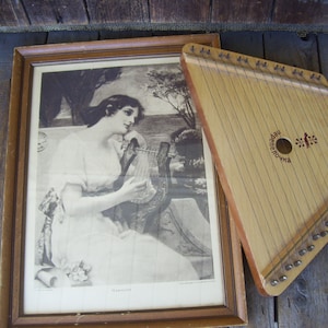 May include: A framed black and white print of a woman playing a harp, with a wooden zither resting on top of the frame. The zither has a label that reads "Народная Цитpa".