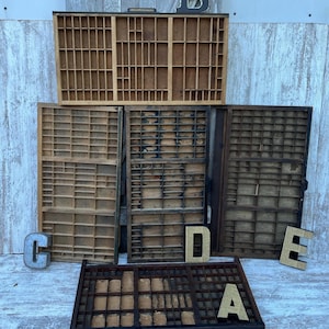 May include: A collection of five rectangular wooden printer's type cases with multiple small compartments, arranged in a stack. The set includes decorative metal letters B, C, D, E, and A placed around the wooden trays on a white surface.