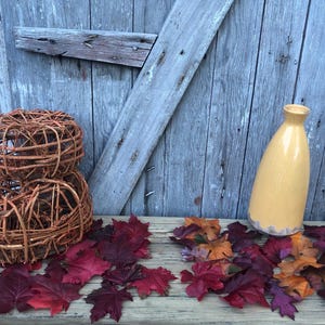 May include: A rustic still life featuring a yellow ceramic vase and two woven wicker spheres. The scene is adorned with colorful fall leaves in shades of red, orange, and purple. The background is a weathered wooden door.
