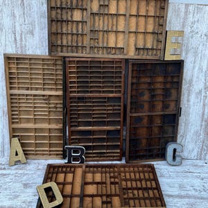 May include: A collection of five rectangular wooden printer trays featuring numerous small compartments of varying sizes. The trays exhibit different wood tones ranging from light to dark brown. Decorative metal letters A, B, C, D, and E are placed.