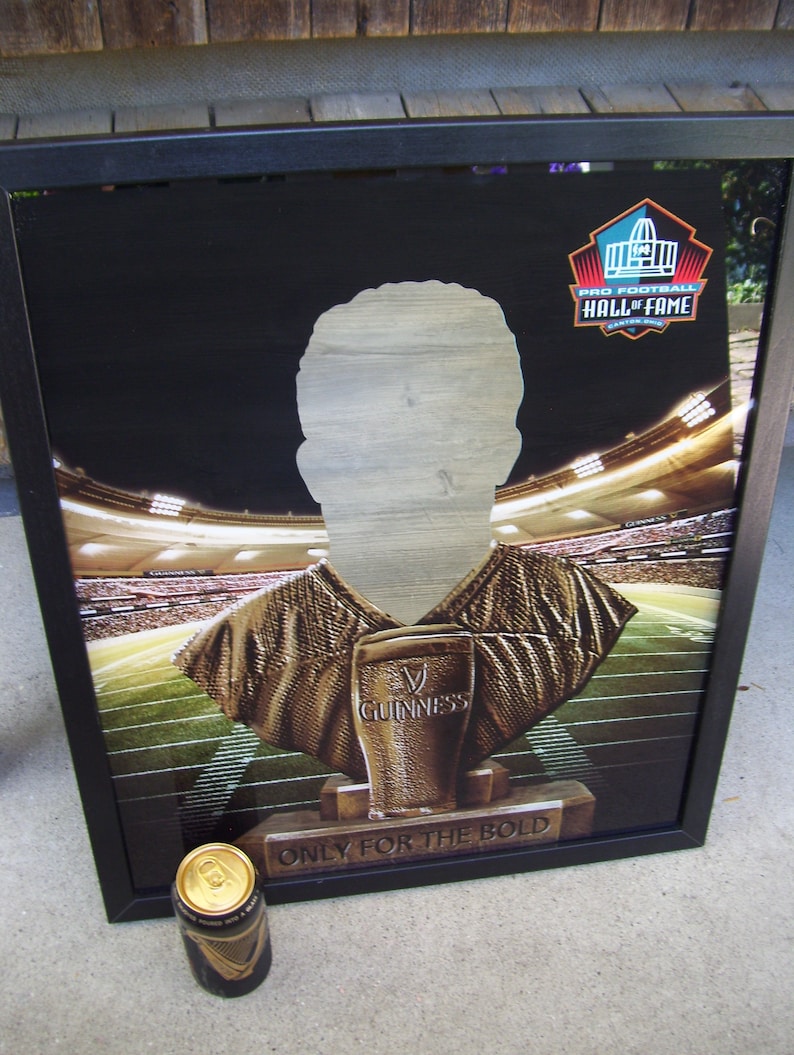 May include: A black framed poster with a bronze bust of a football player wearing a jersey. The bust is holding a glass of Guinness beer. The poster has the text "Pro Football Hall of Fame" and "Only for the Bold".