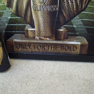 May include: A black and gold can of Guinness beer with the text "Only for the Bold" on a gold base. The can is sitting on a concrete surface.