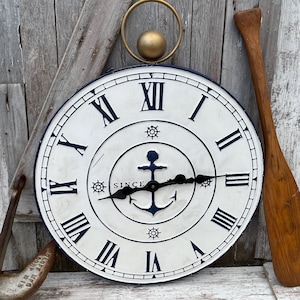 May include: A nautical-themed wall clock with a white face, navy blue Roman numerals, and an anchor design. The clock features a gold-colored ring and sphere at the top. An oar and anchor are visible on the sides. The clock reads "SINCE".