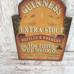May include: A wooden sign with a brown background and gold lettering advertising Guinness Extra Stout. The sign reads "GUINNESS Since 1759 EXTRA STOUT BOTTLED & DRAUGHT the taste of Ireland ARTHUR GUINNESS SON & CO BREWERS & BOTTLERS (DUBLIN) LTD ST. JAMES'S GATE. DUBLIN".