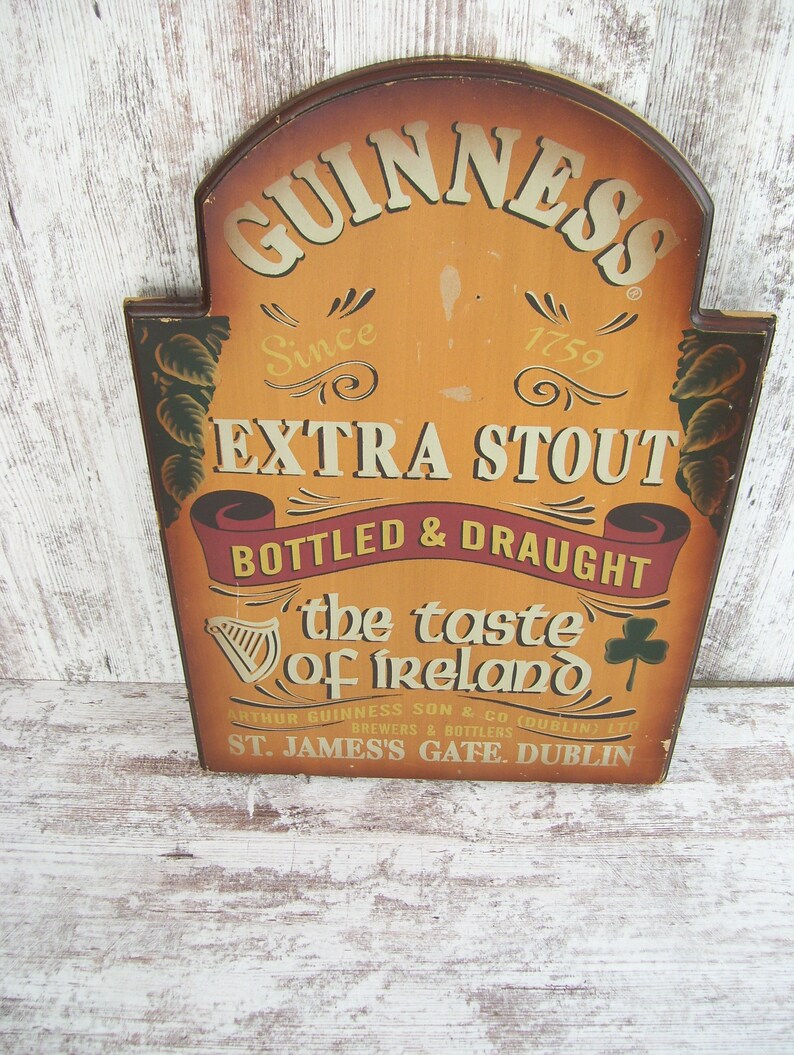 May include: A wooden sign with a brown background and gold lettering advertising Guinness Extra Stout. The sign reads "GUINNESS Since 1759 EXTRA STOUT BOTTLED & DRAUGHT the taste of Ireland ARTHUR GUINNESS SON & CO BREWERS & BOTTLERS (DUBLIN) LTD ST. JAMES'S GATE. DUBLIN".