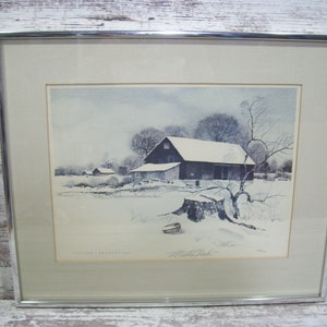 May include: A framed watercolor painting of a snow-covered farmhouse with a barn in the background. The painting is set against a backdrop of a snowy landscape with trees and a single stump in the foreground. The artist's signature is visible in the bottom right corner.