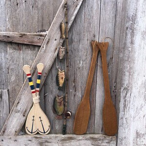 May include: Wooden nautical-themed wall decor. Includes two cream-colored oars with blue anchor designs, a fishing rod with wooden fish, and two wooden oars. The items are arranged against a weathered wooden background.