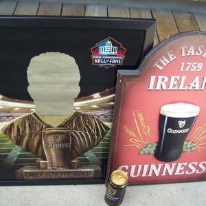 May include: Two wooden signs with a Guinness theme. The first sign is black with a gold football player silhouette and the text "Pro Football Hall of Fame" and "Only for the Bold". The second sign is red with the text "The Taste of Ireland 1759" and a glass of Guinness. Both signs have a brown wood frame.