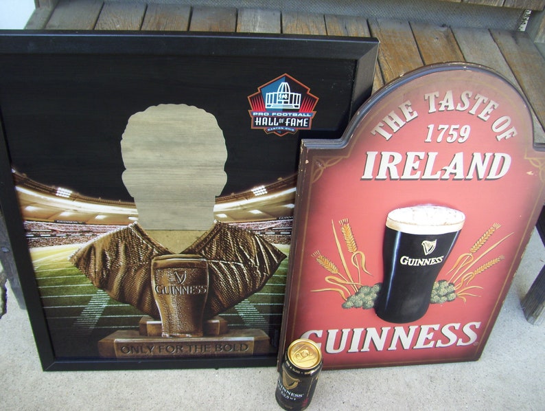 May include: Two wooden signs with a Guinness theme. The first sign is black with a gold football player silhouette and the text "Pro Football Hall of Fame" and "Only for the Bold". The second sign is red with the text "The Taste of Ireland 1759" and a glass of Guinness. Both signs have a brown wood frame.