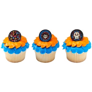 May include: Three decorated cupcakes with orange and blue frosting. Each cupcake has a round, black topper with a different image from the movie Coco. One topper features Miguel, another features a sugar skull, and the last one features a floral pattern.