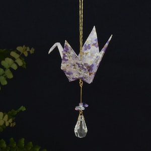 May include: A handcrafted origami crane with a floral pattern in shades of purple and white, suspended by a gold string. A clear crystal teardrop hangs below, adding a touch of elegance. The crane is a decorative item.