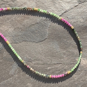 Könnte beinhalten: Eine zarte Halskette mit kleinen, farbenfrohen Perlen. Die Halskette zeigt einen Farbverlauf aus Rosa, Grün und Gold, wodurch ein lebendiges und auffälliges Accessoire entsteht. Die Perlen sind aufgefädelt und bilden ein stilvolles Schmuckstück.