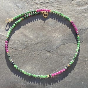 Könnte beinhalten: Eine Halskette mit grünen und pinkfarbenen Edelsteinen. Sie hat einen goldenen Verschluss und kleine goldene Perlen. Die runden Edelsteine sind gleichmäßig angeordnet und bilden ein zartes und farbenfrohes Accessoire.