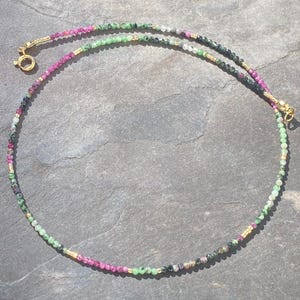Könnte beinhalten: Eine zarte Perlenkette mit kleinen, facettierten, mehrfarbigen Edelsteinperlen in Rosa-, Grün- und Dunkelgrautönen, durchsetzt mit winzigen goldfarbenen Metallzwischenperlen. Die Kette ist mit einem runden, goldfarbenen Federringverschluss versehen.