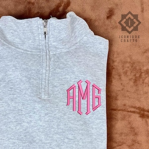 May include: A light gray quarter-zip sweatshirt with a pink monogram of the letters "AMG" embroidered on the chest. The sweatshirt has a high collar and a silver zipper. The logo "ICONIQUE CRAFTS" is in the upper right corner.