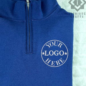 May include: A royal blue quarter-zip pullover featuring a silver-toned metal zipper at the collar. On the left chest, a circular white graphic contains the text YOUR LOGO HERE with a star on each side of the word LOGO.
