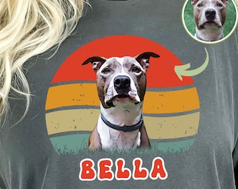 May include: A gray t-shirt with a retro-style sunset graphic featuring a dog with the name "BELLA" printed below. The dog is a white and brown pit bull terrier. The background is a grunge-style design.